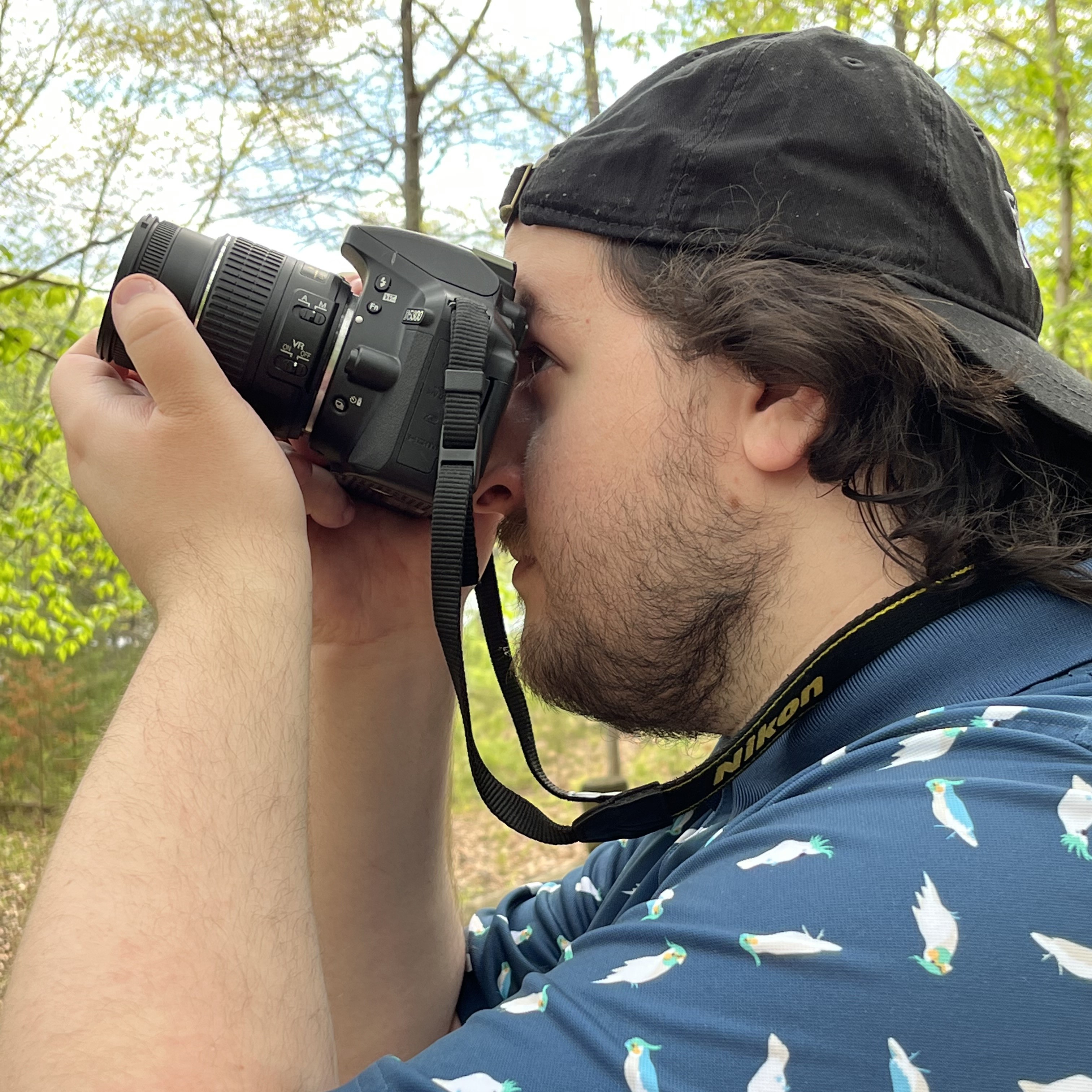 Mężczyzna w czarnej czapce i niebieskiej koszulce z motywem ptaków, trzymający aparat fotograficzny Nikon i patrzący przez wizjer, na tle zielonych drzew.